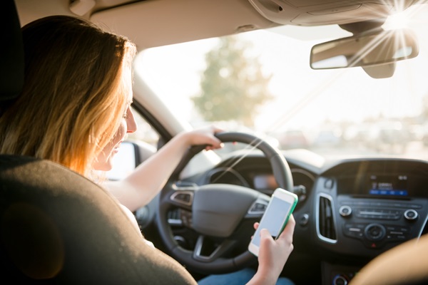 A young woman with blonde hair is sitting in the driver's seat of a car, holding a smartphone in her right hand and looking at the screen. Her left hand is on the steering wheel.